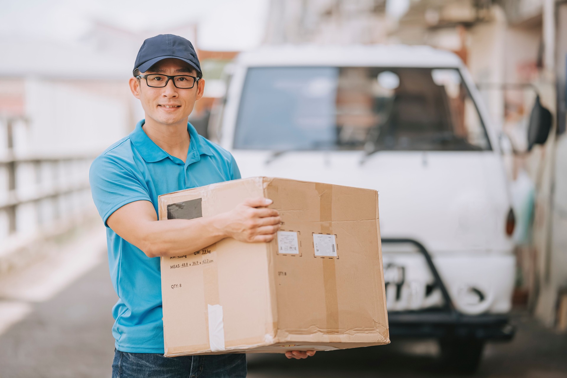 asian chinese mature male delivery person holding cardboard box looking at camera smiling in front of delivery van